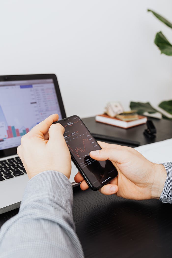 Person analyzing stock market data on smartphone, with laptop in background.